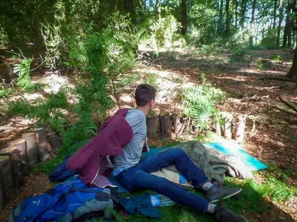 Person sitzt auf einer Unterlage im Wald, umgeben von Rucksack und Ausrüstung