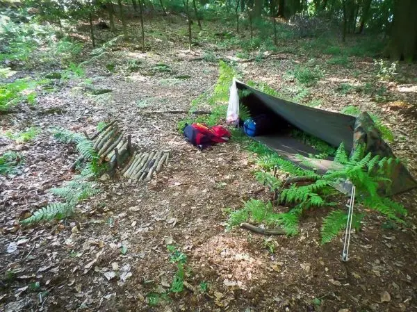 Tarp-Notunterkunft mit Rucksack und Holzstapel im Wald