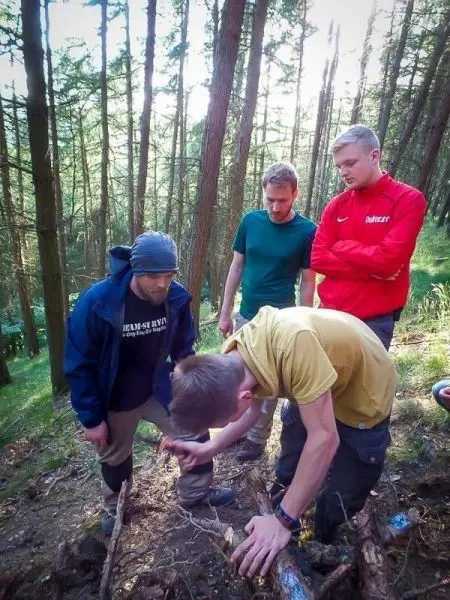 Vier Personen beobachten einen Mann, der am Boden mit Holzstücken arbeitet