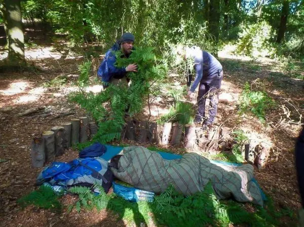 Zwei Personen bauen eine Notunterkunft aus Ästen und Blättern im Wald