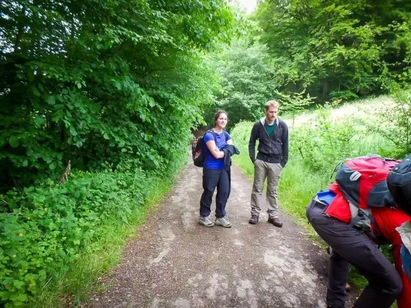 Zwei Personen stehen auf einem Waldweg, eine Person beugt sich zu einem Rucksack