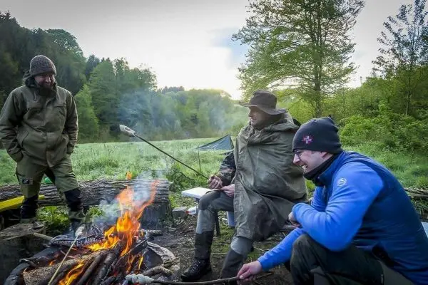 Drei Personen sitzen um ein Feuer und rösten etwas über den Flammen