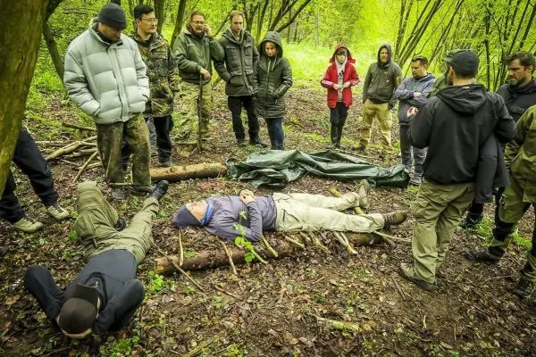 Gruppe beobachtet zwei Personen auf improvisierten Tragen aus Ästen im Wald
