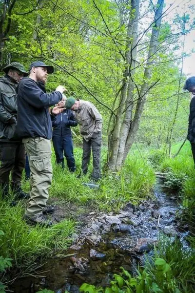 Gruppe betrachtet einen kleinen Bachlauf zwischen Bäumen und hohem Gras