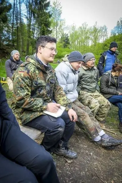 Gruppe sitzt auf Holzstühlen im Wald, einige halten Notizen in den Händen