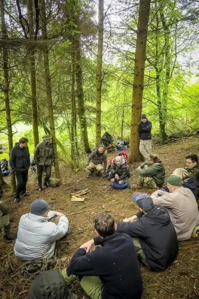 Gruppe sitzt im Wald auf dem Boden und hört einem Vortrag zu