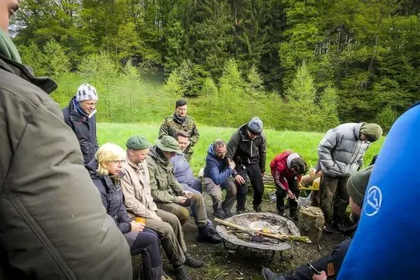 Gruppe sitzt um ein Feuer in einer Freiluftumgebung, einige arbeiten mit Holz