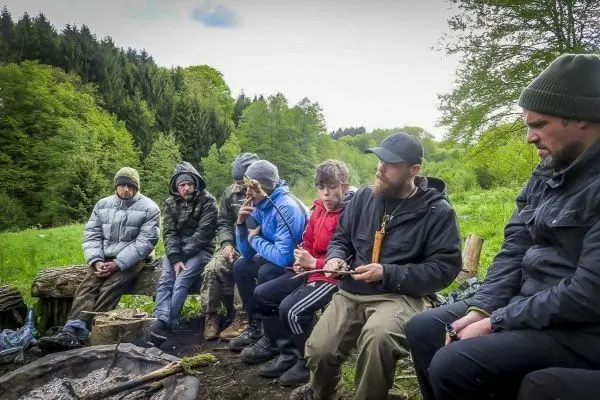 Gruppe sitzt um ein kleines Bodenfeuer und arbeitet an Holzprojekten