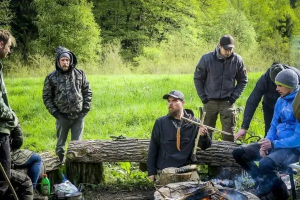 Gruppe um ein Lagerfeuer versammelt, Holzbank im Vordergrund, einige Personen halten Werkzeuge