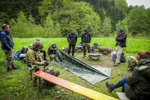 Gruppe um Tarp-Notunterkunft versammelt, einige sitzen auf Bänken, andere stehen