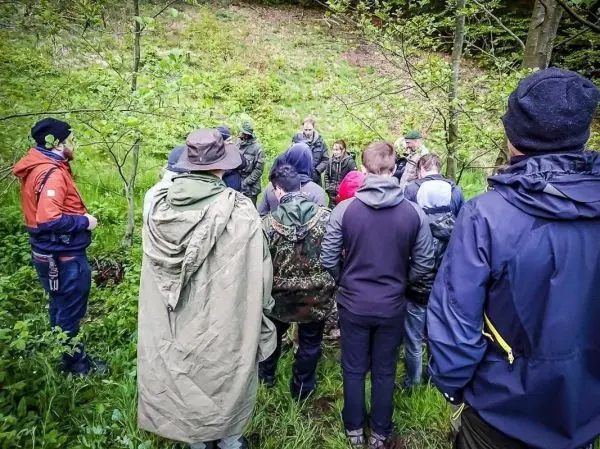 Gruppe von Personen hört einem Vortrag im Wald zu, umgeben von Bäumen und Gras