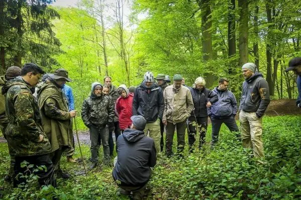 Gruppe von Personen steht im Wald und beobachtet einen knienden Trainer