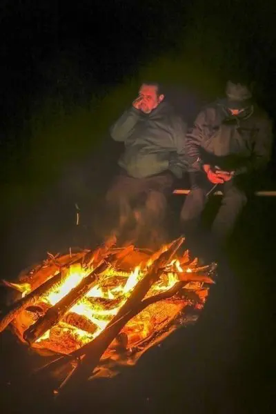 Kleine Flamme brennt zwischen Holzstücken, zwei Personen sitzen daneben