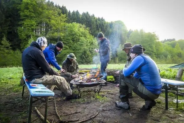 Kleines Bodenfeuer mit Rauch, mehrere Personen sitzen und stehen um das Feuer