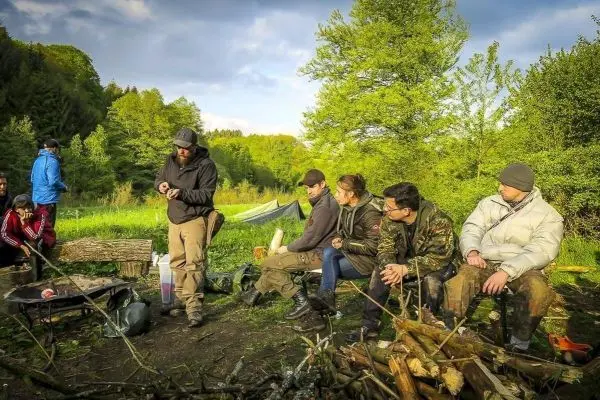 Lagerfeuer brennt vor sitzenden Personen, Holzstapel und Tarp-Notunterkunft sichtbar