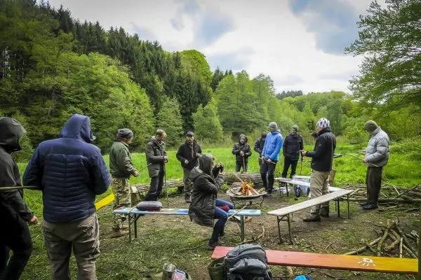 Lagerfeuer in einer Waldlichtung, umgeben von mehreren Personen und Bänken