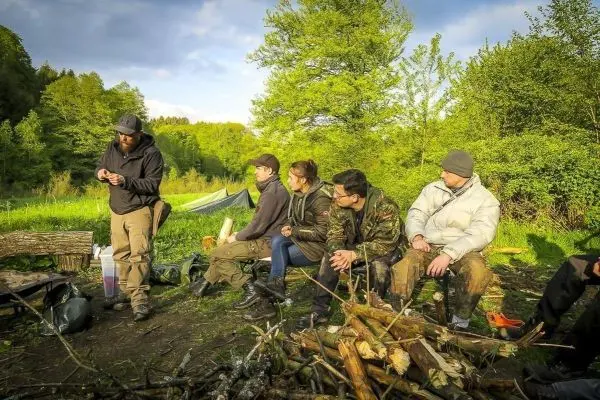 Lagerfeuer mit Holzstapel, Teilnehmer sitzen auf Baumstämmen und beobachten