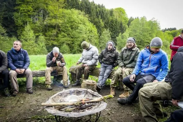 Lagerfeuer mit Kochtopf auf einem Metallgestell, umgeben von mehreren Personen auf Baumstämmen