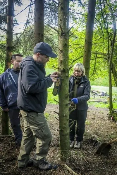 Mann befestigt ein Werkzeug an einem Baumstamm, während zwei Personen beobachten