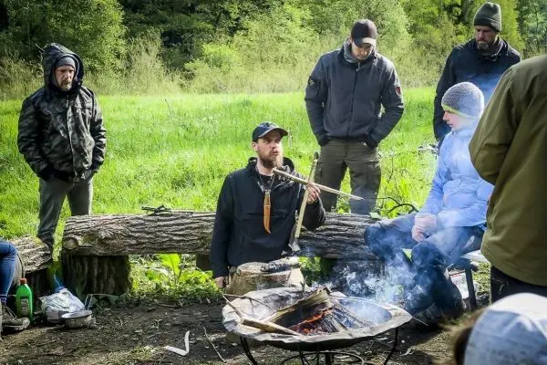 Mann demonstriert Feuerbohren über einem kleinen Feuer, Zuschauer beobachten aufmerksam