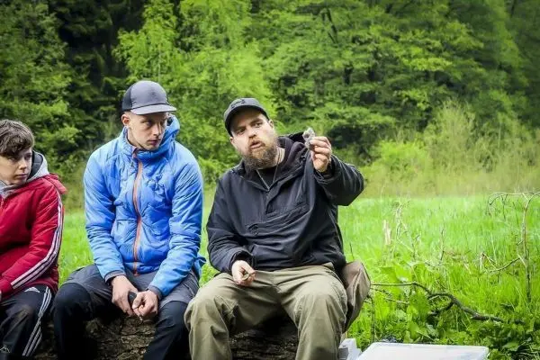 Mann erklärt etwas und hält einen Gegenstand in der Hand, zwei Zuhörer sitzen daneben