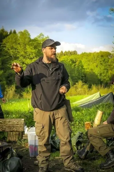 Mann erklärt Techniken im Freien, Holz und Ausrüstung um ihn herum sichtbar
