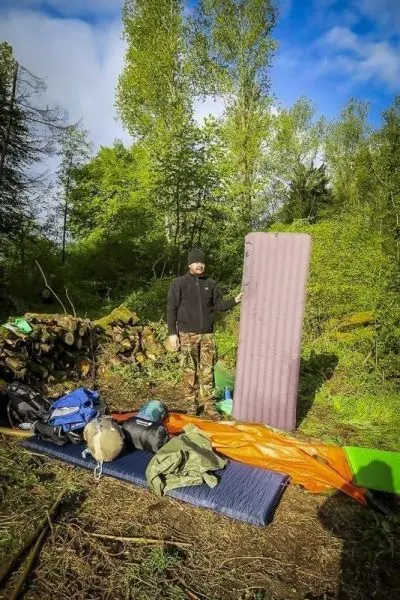Mann hält eine Isomatte in einem Waldgebiet, Rucksäcke und Ausrüstung liegen am Boden
