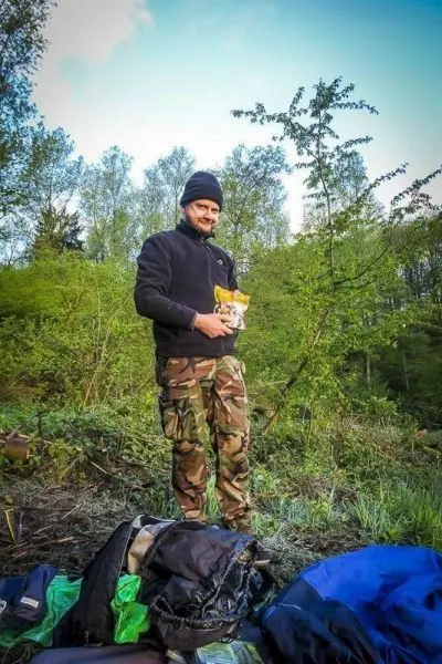 Mann hält eine Packung Nahrung in der Hand, umgeben von Rucksack und Natur