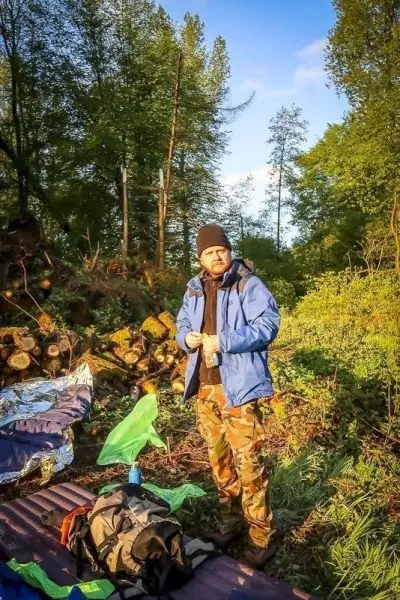 Mann in blauer Jacke und Mütze steht neben Rucksack und Schlafunterlage im Wald
