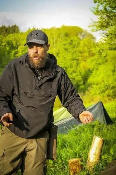 Mann in schwarzer Jacke erklärt etwas vor einem Tarp und Holzstücken