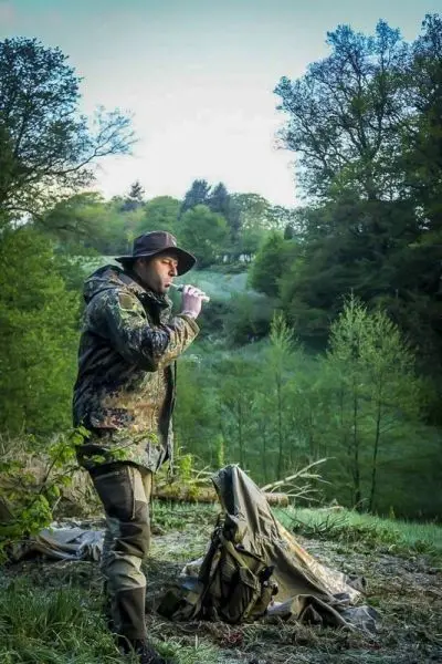 Mann in Tarnkleidung steht neben einer Tarp-Notunterkunft und trinkt aus einer Flasche