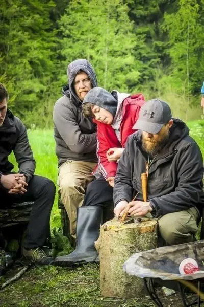Mann schnitzt mit einem Messer an einem Holzstück, während andere zuschauen