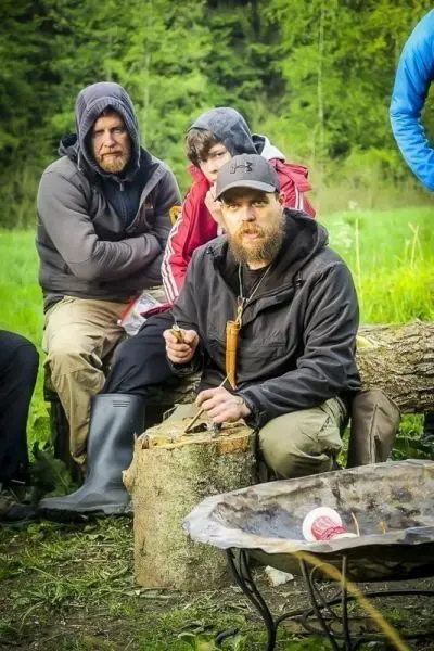 Mann schnitzt mit einem Messer auf einem Baumstamm, im Hintergrund sitzen weitere Personen