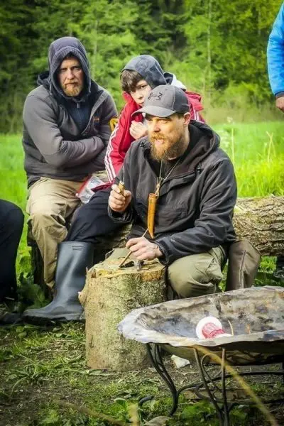 Mann schnitzt mit einem Messer auf einem Baumstumpf, während andere zuschauen