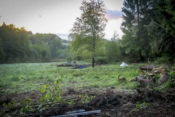 Mann steht in einer Wiese mit Zelten und Holzstapeln im Hintergrund