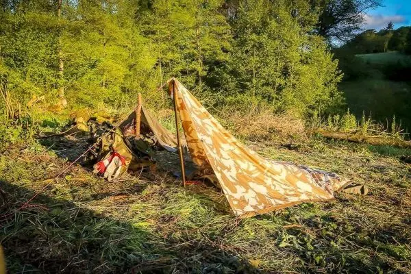 Tarp-Notunterkunft aus Camouflage-Stoff mit Rucksack daneben auf dem Boden
