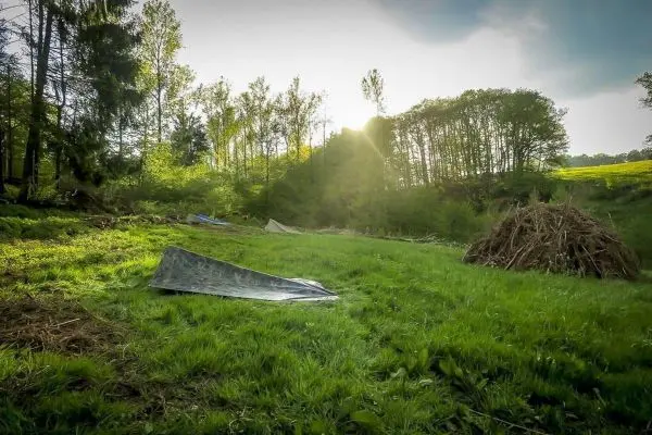 Tarp-Notunterkunft liegt auf grünem Gras, im Hintergrund Holzstapel und Bäume