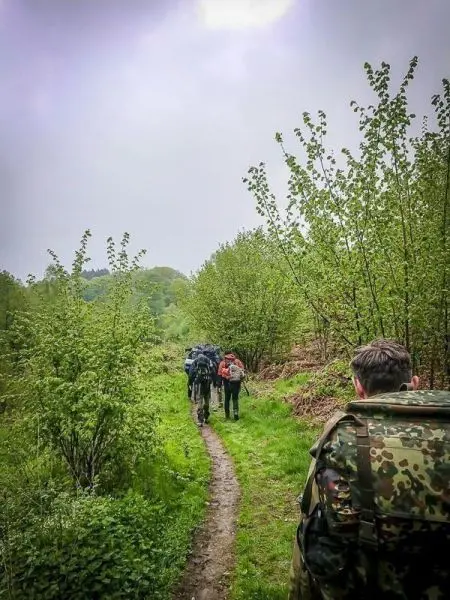 Wandernde Personen mit Rucksäcken auf einem schmalen Pfad zwischen Bäumen