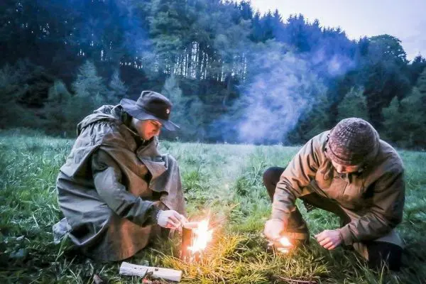 Zunder glimmt auf dem Boden, während zwei Personen Feuer entfachen
