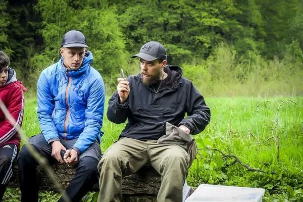 Zwei Männer sitzen auf einem Baumstamm und hören einem dritten Mann zu, der spricht