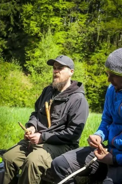 Zwei Personen sitzen im Freien und bearbeiten Holzstücke mit Messern