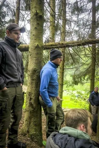Zwei Personen stehen an einem Baumstamm im Wald, eine weitere Person sitzt im Vordergrund
