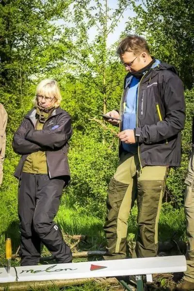 Zwei Personen stehen auf einer Holzbank im Freien, eine schaut nachdenklich, die andere nutzt ein Smartphone