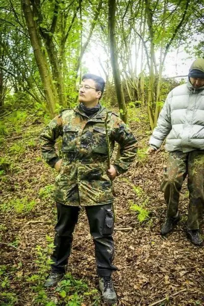 Zwei Personen stehen im Wald, eine trägt eine Tarnjacke und die andere eine graue Jacke