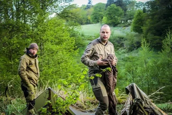 Zwei Personen stehen in einer bewaldeten Umgebung neben einer Tarp-Notunterkunft