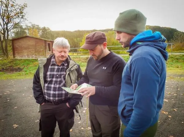 Drei Personen betrachten eine Karte im Freien, umgeben von Bäumen und einer Holzhütte