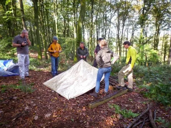Gruppe baut Tarp-Notunterkunft im Wald auf, umgeben von Bäumen und Laub