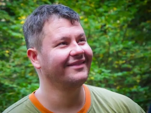 Lächelnde Person mit kurzem Haar vor grüner Waldkulisse