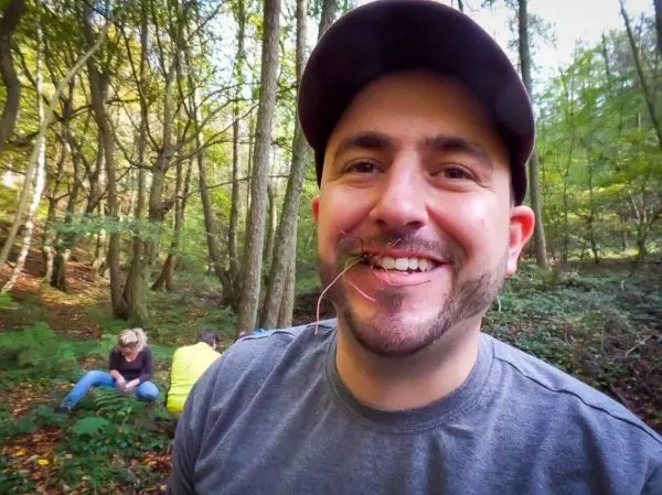 Lächelnder Mann mit Pflanze im Mund, im Hintergrund zwei Personen im Wald