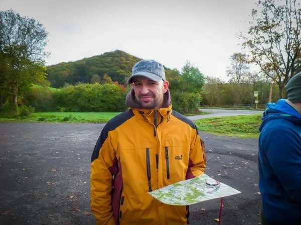 Mann hält eine Karte und Kompass in der Hand, im Hintergrund Hügel und Bäume